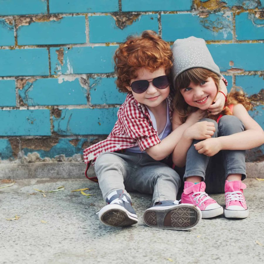 Niños sonrientes con gafas y gorro en la calle junto a pared de ladrillos azul.